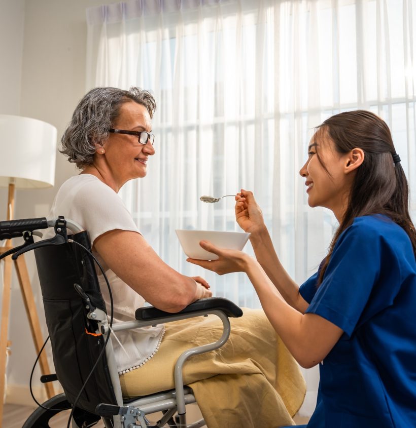 Asian caregiver nurse serve food to Caucasian senior older woman at home. Attractive specialist carer women support and taking care of older elderly mature grandmother in living room in nursing house.