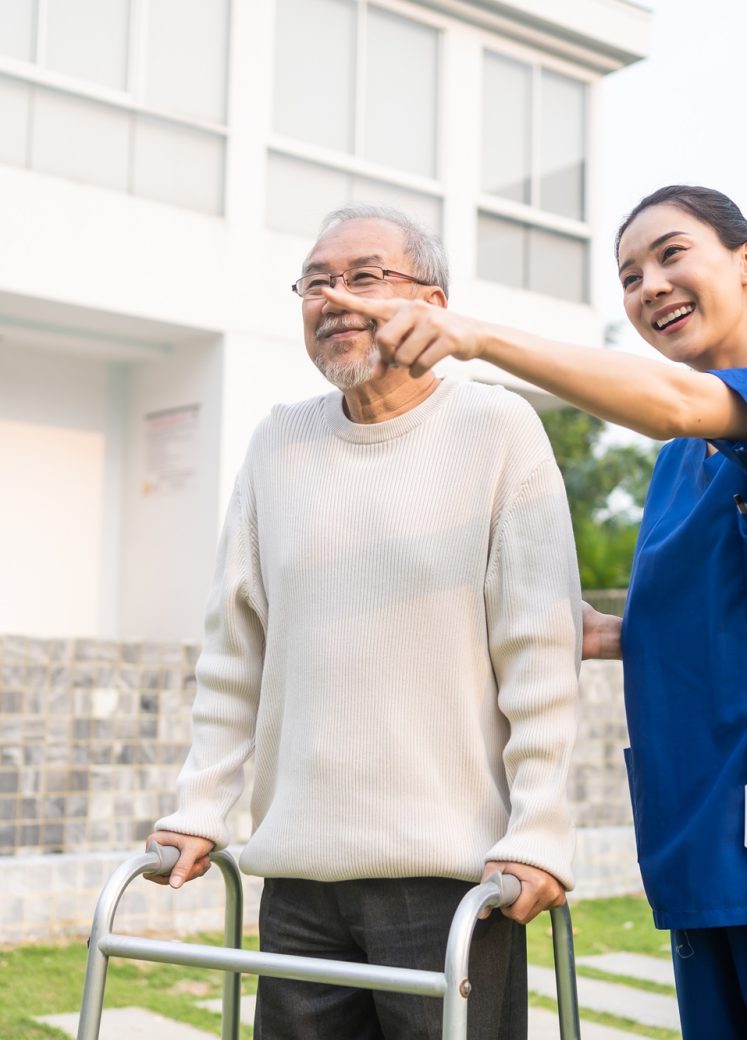 Asian young caregiver nurse support senior older male walking outdoors. Specialist girl doctor help and take care of elderly mature man patient doing physical therapy in public park at nursing home.